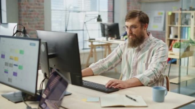 年轻的高加索大胡子男子的肖像，戴着眼镜，在明亮宽敞的办公室里坐着在电脑上工作时微笑。男性法律顾问在键