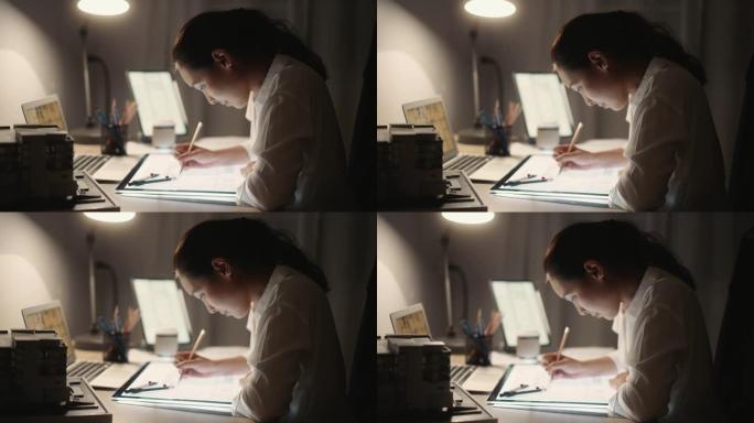 建筑师女性拿着铅笔在夜间使用笔记本电脑和蓝图进行建筑计划