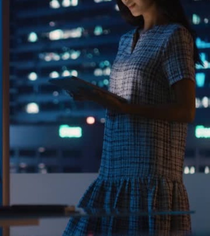 使用平板电脑的金融分析师的垂直肖像，站在窗户旁边，沿着城市的街道，摩天大楼上有霓虹灯。在投资银行城市