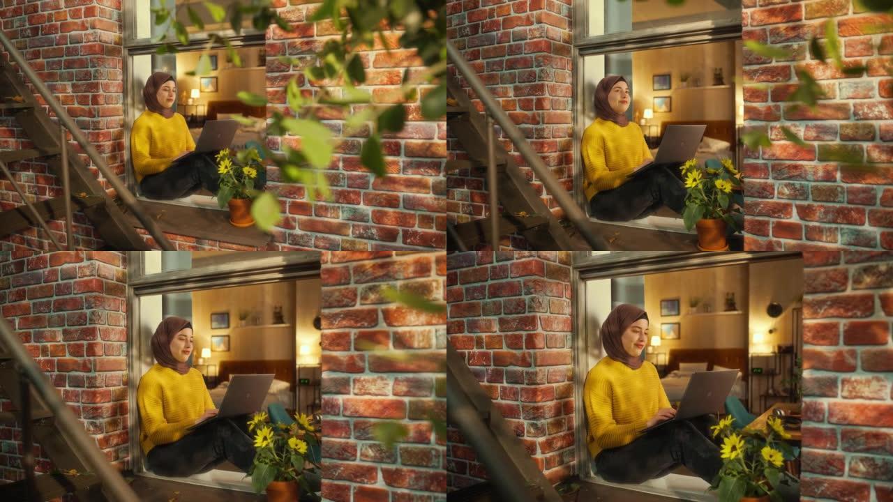 一位年轻的阿拉伯妇女坐在多层砖房的窗台上时使用笔记本电脑的肖像。在家工作时，有动力的女性微笑和快乐。