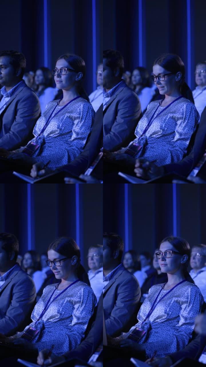 垂直屏幕: 女人坐在商务会议上拥挤的观众中。使用笔记本电脑的女性代表。经理观看关于发展市场的鼓舞人心