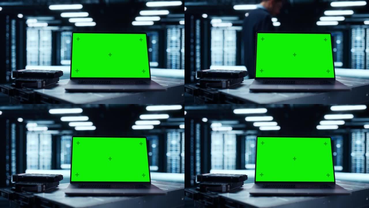 带有绿屏色度键模拟显示屏的笔记本电脑放在桌子上。在后台数据中心，服务器机架行和IT工程师在工作。静态