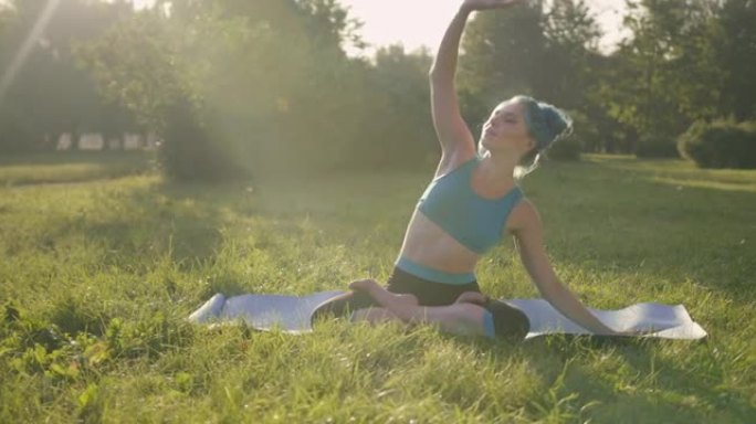 迷人的年轻女子，蓝色的头发，穿着蓝色的瑜伽服装，在公园的草地上做瑜伽，在日出或日落时，在瑜伽垫上的莲