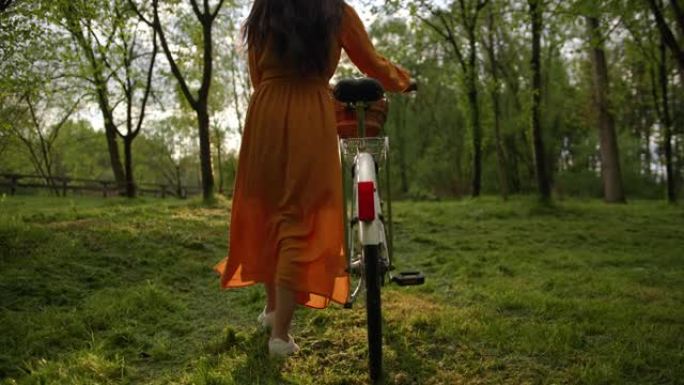 中等，跟踪镜头，一个无法辨认的黑发穿着橙色连衣裙，在森林里骑自行车走向日落，慢动作