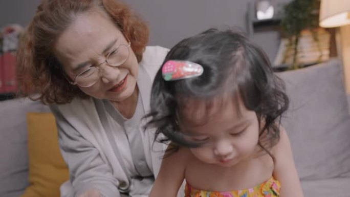 亚洲老人微笑的祖母在家里的客厅里和孙子或孙女一起玩，老年女性开心的同时照顾她的小可爱孩子玩数字平板电