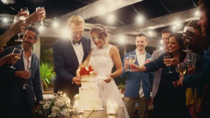美丽的新娘和新郎在晚宴上与多民族朋友庆祝婚礼。已婚夫妇站在餐桌上，亲吻并切婚礼蛋糕。