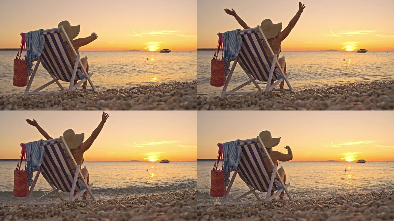 SLO MO欣喜若狂的女人在日落时坐在海滩上的椅子上时将双臂举到天空