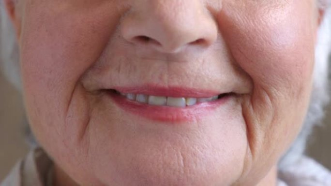 微笑的高级女性在穿着粉红色唇膏和化妆时的嘴唇特写。一位皱着皱纹的老年女士的脸部和嘴巴细节，露出牙齿和