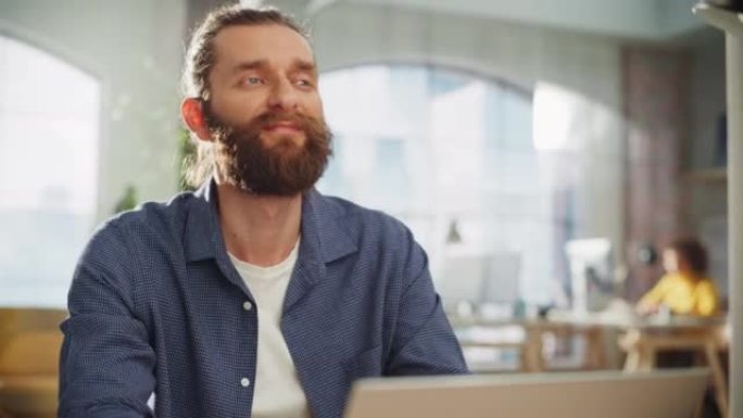 特写一位英俊的长发大胡子经理坐在创意办公室的办公桌前的肖像。时尚的男人在营销公司使用笔记本电脑。撰写