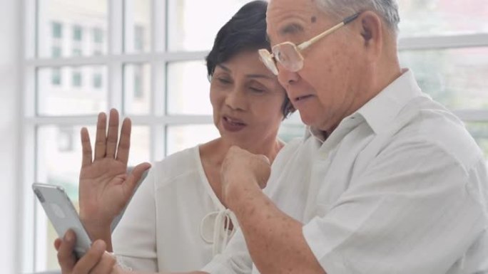 60-65岁的亚洲老年人夫妇在使用智能手机和视频会议技术进行社交距离时，在家中通过视频会议与医护人员