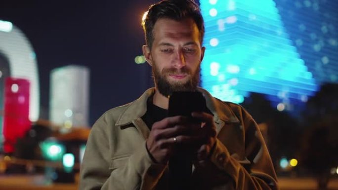 一个大胡子的人在黑暗的城市街道上通过电话阅读新闻信息。霓虹灯晚楼外面有智能手机的人。带有电子显示屏和