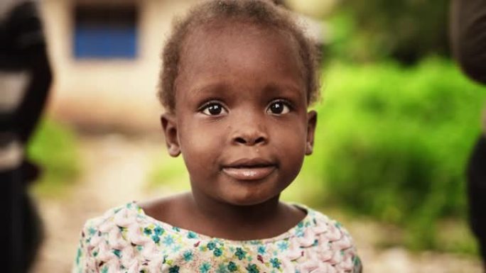一个可爱的非洲小托德勒看着相机的特写肖像，背景模糊的人在移动。充满纯真和喜悦的黑人孩子，代表着对美好