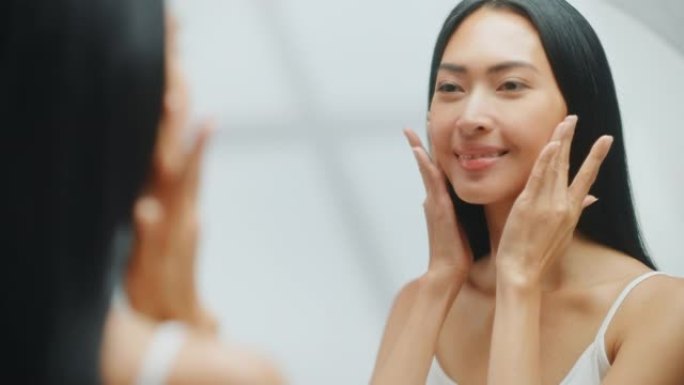 美丽的亚洲女人的肖像轻轻地涂面霜看着浴室的镜子。年轻的成年女性用天然化妆品护肤品让她的皮肤柔软、光滑