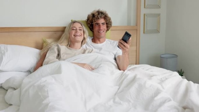 夫妻在床上在卧室看电视，一起观看流媒体电影或电视节目，一起享受早晨的美好时光。关系，男人和女人在家看