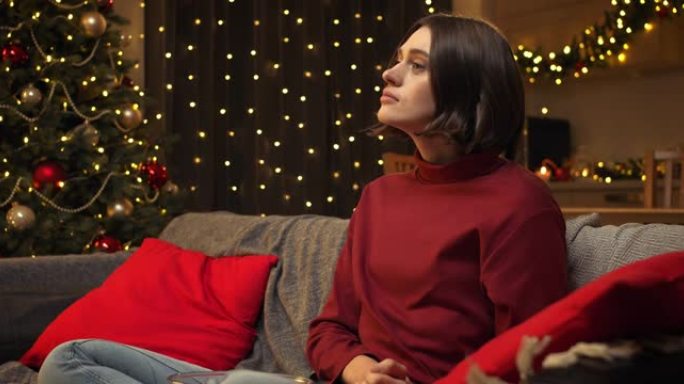 一个穿着舒适的红色毛衣的漂亮棕色头发的女人看着电视剧，担心角色，坐在圣诞节装饰的房间里