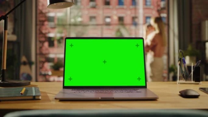 笔记本电脑显示屏，模拟绿屏站在客厅的木桌上。家里或创意阁楼办公室的色度钥匙监视器，背景是女性向外看。