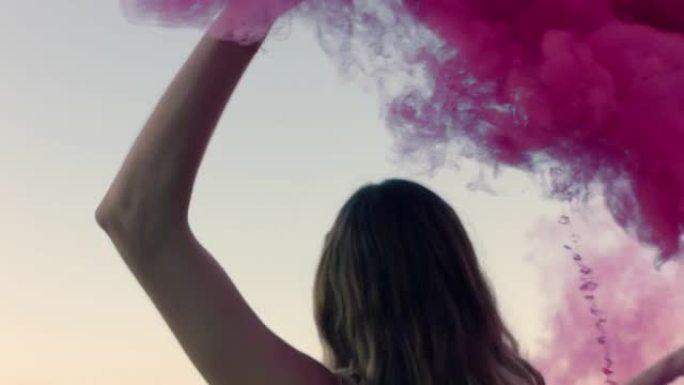 美丽的女人挥舞着粉红色的烟雾弹在日出时在海滩上跳舞，庆祝创意自由