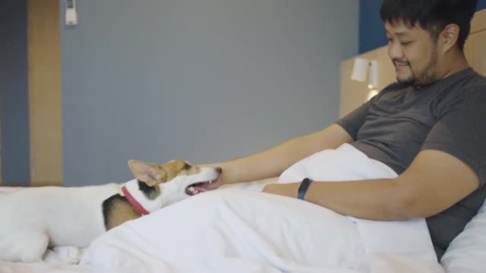SLO MO: 男人坐在床上，与他的狗保持联系，他的狗在酒店度假期间幸福地躺在他身上。