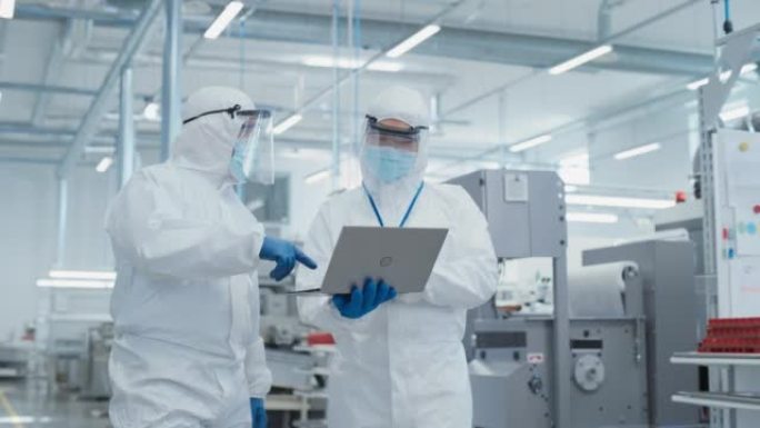 两名科学家穿着无菌工作服和口罩站在重工业工厂，使用笔记本电脑。检查工业机器设置和配置生产功能。