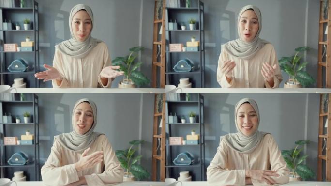 迷人的年轻亚洲穆斯林女商人戴着头巾，随意坐在桌子上看着相机远程工作视频在线通话在家里客厅的笔记本电脑