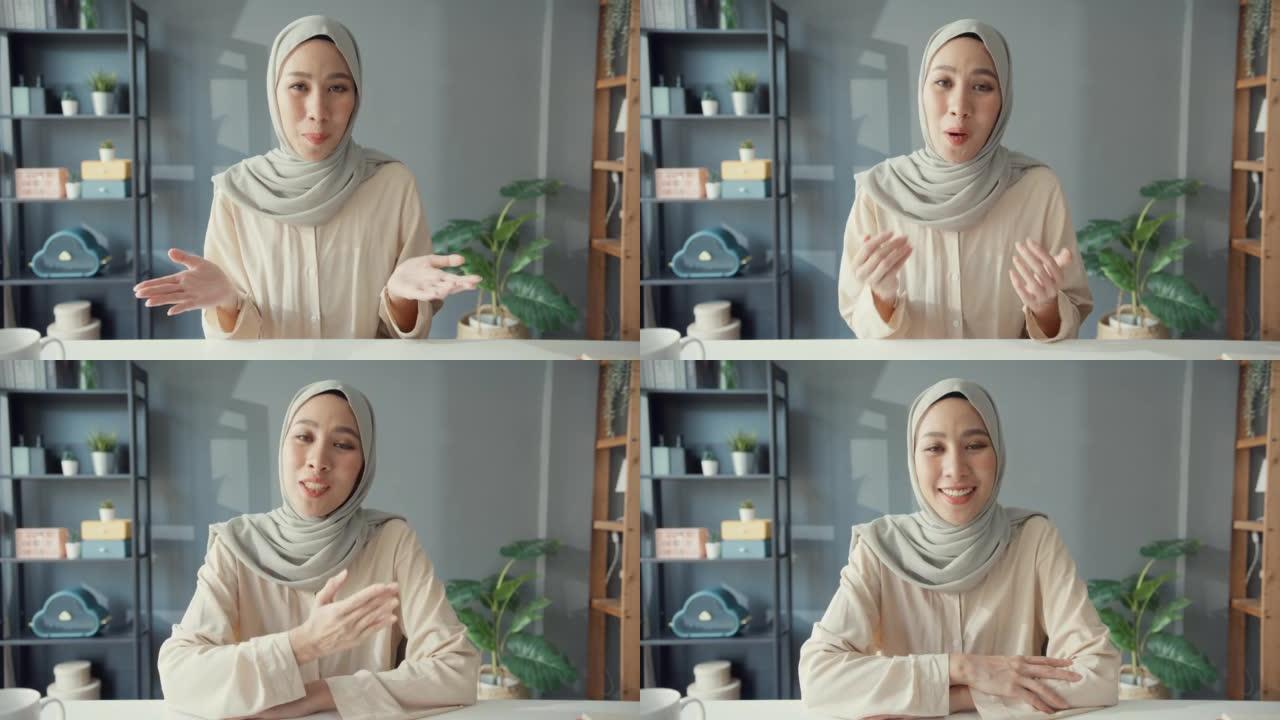 迷人的年轻亚洲穆斯林女商人戴着头巾，随意坐在桌子上看着相机远程工作视频在线通话在家里客厅的笔记本电脑