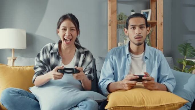 快乐的亚洲年轻夫妇坐在沙发上使用操纵杆控制器玩视频游戏在客厅一起度过快乐的时光。
