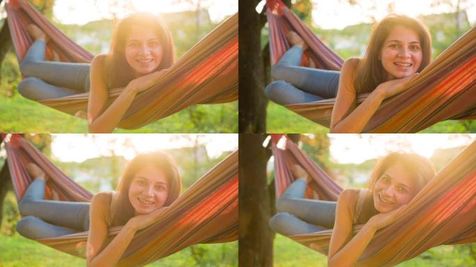 快乐的女人在郁郁葱葱的绿色植物中的吊床上摇摆，对着镜头微笑