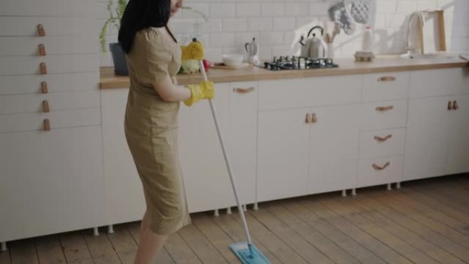 年轻女子用现代拖把洗地板，在家厨房跳舞的慢动作肖像。