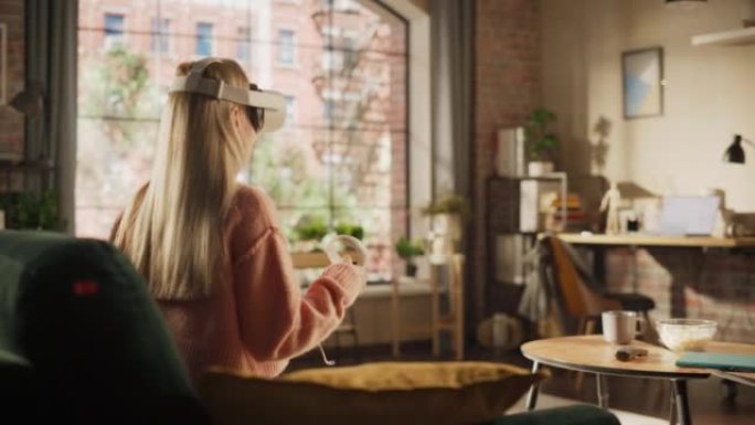 金色头发的女性在家中使用虚拟现实耳机和控制器。有创造力的女人坐在客厅的沙发上，玩VR视频游戏或在特殊