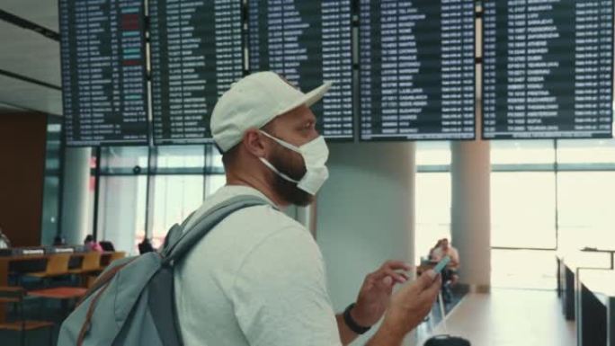 年轻的白人游客戴着口罩，在机场航站楼使用智能手机，检查航班时刻表，走开了。