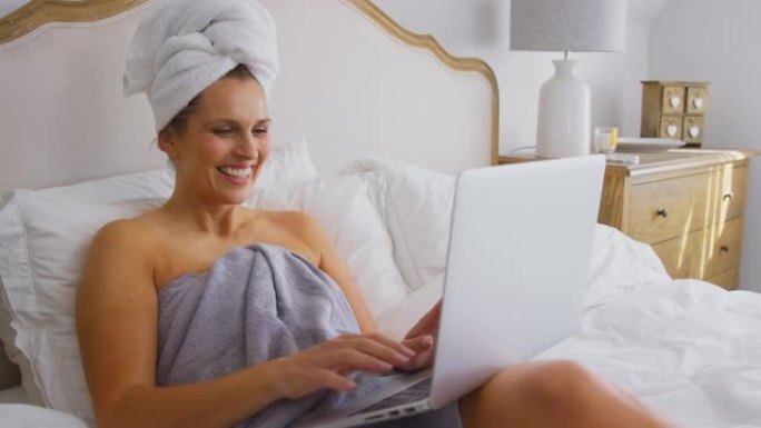 成熟的女人躺在床上，在家穿着毛巾在笔记本电脑上进行视频通话
