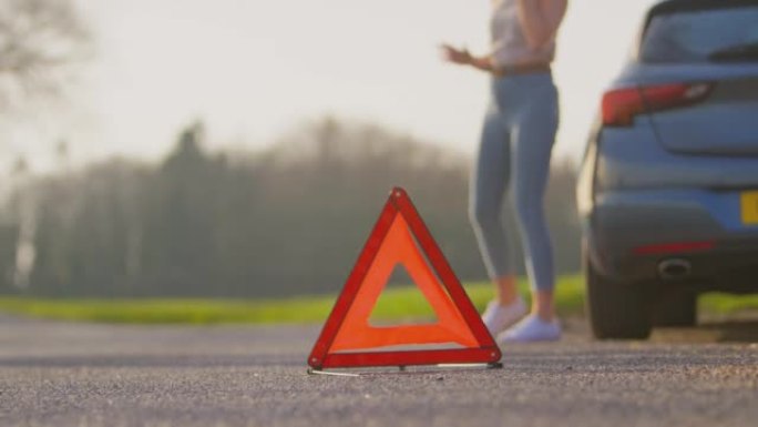 前景中的乡村道路上的危险警告标志，女人站在引擎盖上的抛锚汽车旁边，用手机呼救-慢动作拍摄