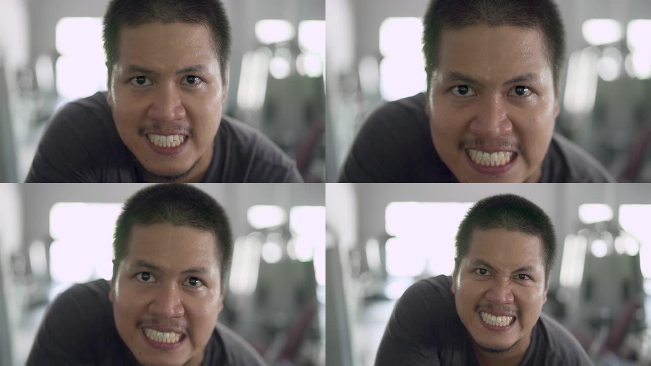 慢动作光头亚洲男子在健身房展示自己的力量，脸上流汗看着相机