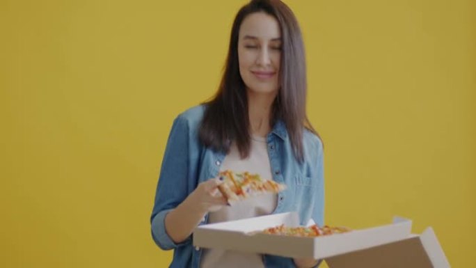 饥饿的年轻女士打开盒子的肖像看着比萨饼，在黄色背景上微笑着吃切片