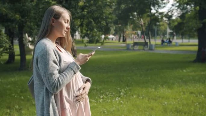 美丽孕妇走在城市公园用智能手机录制音频信息的侧视图