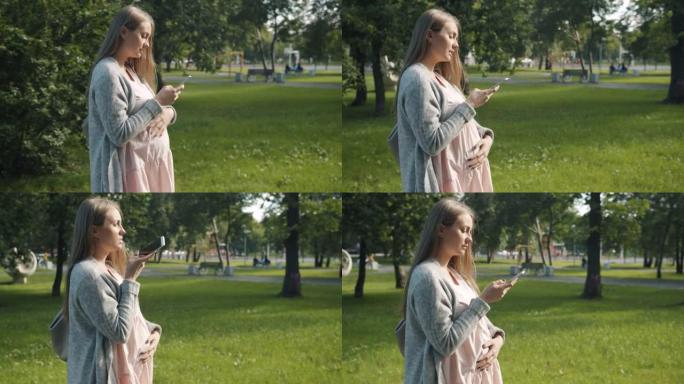美丽孕妇走在城市公园用智能手机录制音频信息的侧视图