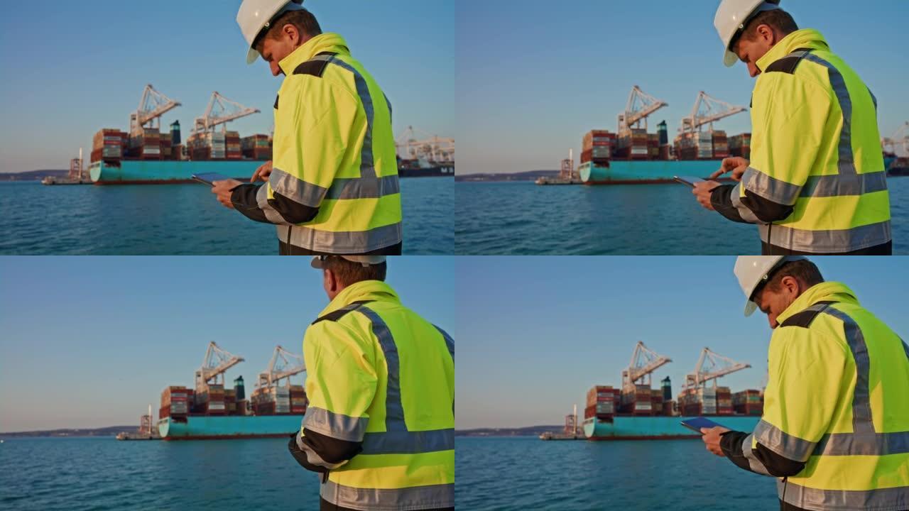 工程师，戴着头盔和警告背心的人使用他的数字平板电脑，站在港口时看着集装箱船