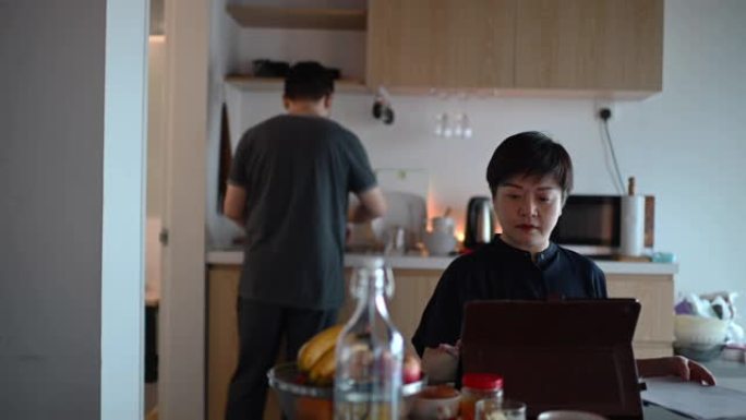 一名亚洲中国中年妇女在早餐期间在家在餐桌上工作，使用数字平板电脑和文件，同时丈夫洗碗