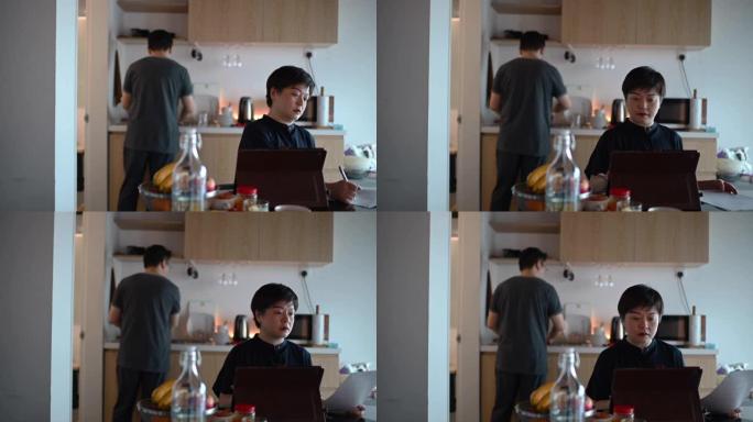 一名亚洲中国中年妇女在早餐期间在家在餐桌上工作，使用数字平板电脑和文件，同时丈夫洗碗