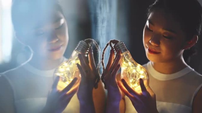一个美丽的亚洲中国少女看着并拿着一个带发光二极管灯串的灯泡，用镜子反射微笑