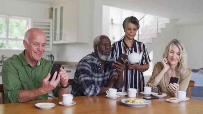 两对不同的老年夫妇坐在桌子旁，在家中使用智能手机喝茶