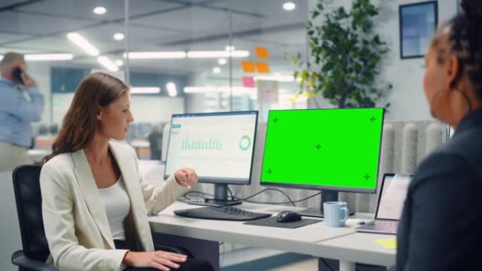 两名女同事在现代商务办公室使用绿屏模拟显示的计算机上工作时，彼此亲切地交谈，笑着笑。经理和员工讨论一
