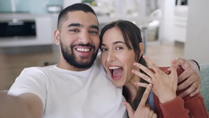 年轻兴奋的夫妇分享订婚消息的肖像，在视频通话中显示铃声。年轻的有影响力的夫妇在家里的休息室沙发上自拍