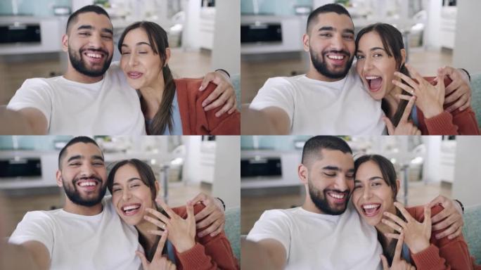 年轻兴奋的夫妇分享订婚消息的肖像，在视频通话中显示铃声。年轻的有影响力的夫妇在家里的休息室沙发上自拍