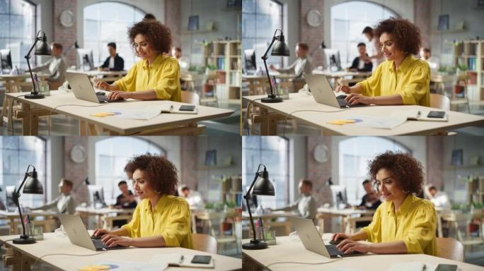 在现代繁忙的办公室里，混血儿富有成效的年轻女子在电脑上工作的肖像。女队在完成一项重要任务后面带微笑。