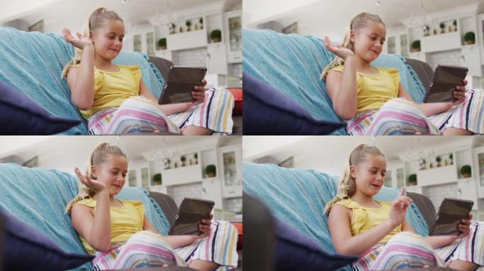 快乐的高加索女孩在家，坐在沙发上使用平板电脑进行视频通话并挥手
