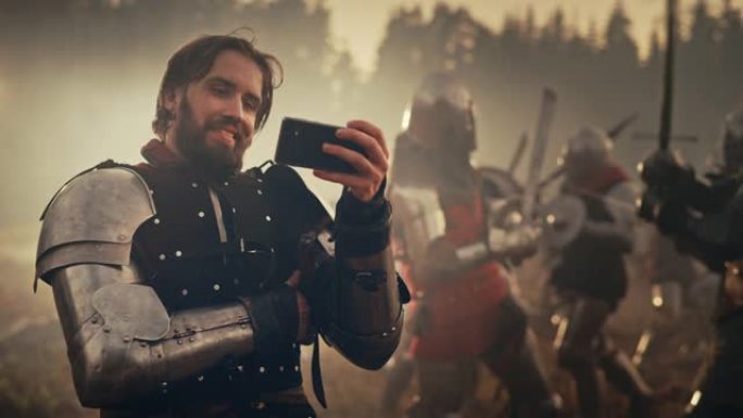 英俊的中世纪骑士在战场上微笑着用智能手机。装甲战士看在线游戏，点播搞笑视频。在背景中，战争是愤怒的