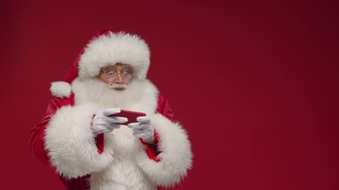 一个开朗的圣诞老人热情地在红色的智能手机上玩耍，然后他把它藏在背后，被宠爱，站在红色的背景下