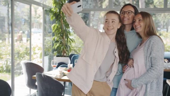 快乐的朋友在咖啡馆与孕妇触摸腹部自拍为智能手机相机摆姿势