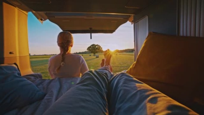 在阳光明媚的日子里，一个男人躺在机舱里，女人坐在停在乡下某个地方的露营者前面